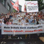  150 Personen stehen auf einer Treppe. Sie halten Kartonschilder und Fahnen in den Händen. Die vorderste Reihe hält ein Transparent, auf dem «Mehr Schutz dank starkem GAV: Unsere Zukunft, unsere Rechte.» steht.