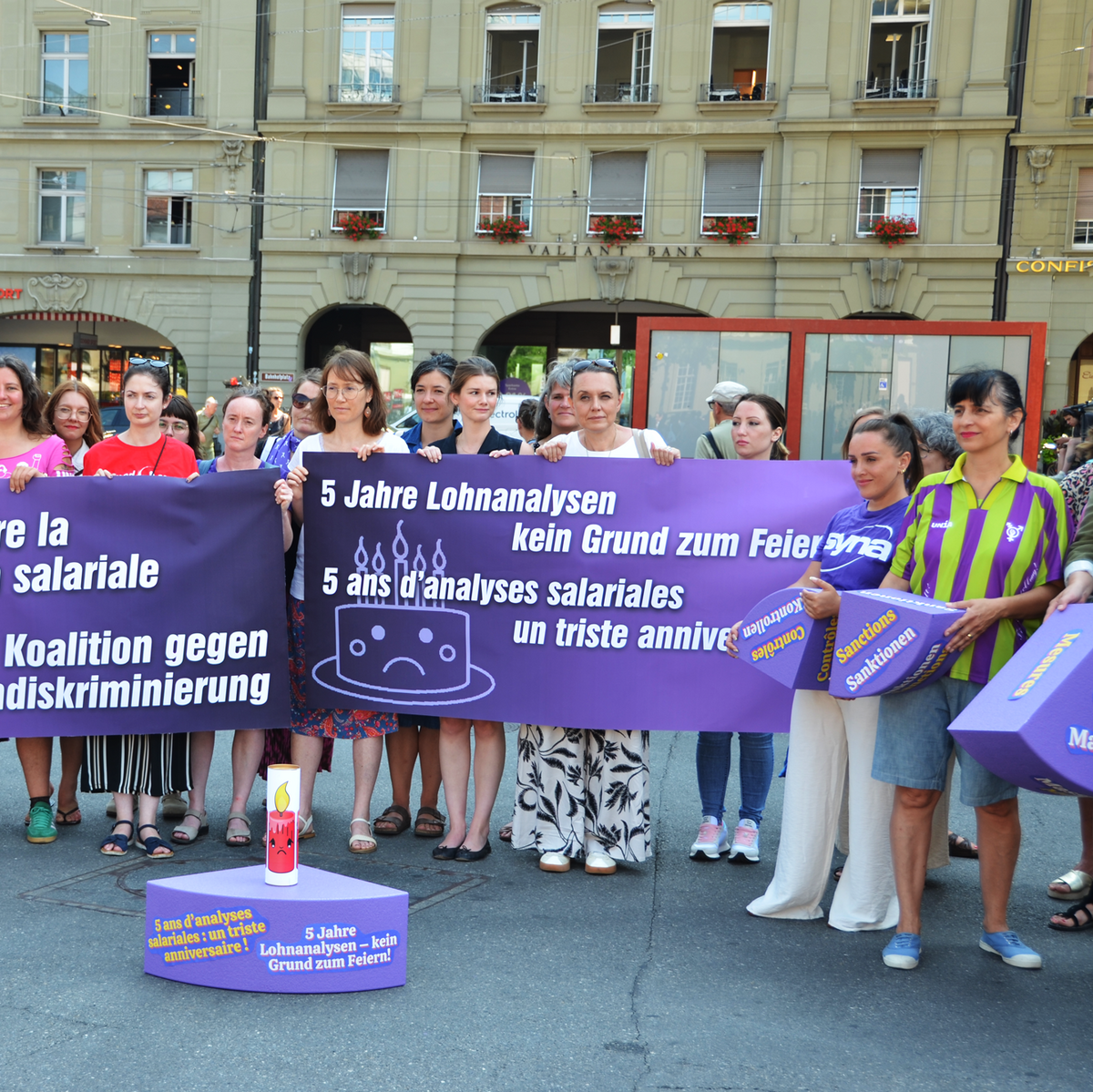 Knapp 30 Frauen aus verschiedenen progressiven Organisationen protestieren in Bern auf dem Bahnhofplatz. Sie stehen vor einem riesigen violetten Kuchenstück aus Styropor - eines von einem ganzen Kuchen, dessen einzelne Stücke die jeweiligen Mängel des Gleichstellungsgesetzes symbolisieren. 