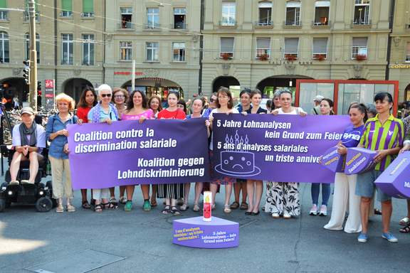 Una trentina di donne appartenenti a diverse organizzazioni progressiste protestano sulla Piazza federale di Berna. Sono in piedi davanti a una gigantesca torta di polistirolo viola. I singoli pezzi di torta simboleggiano le rispettive lacune della legge sulla parità di genere.