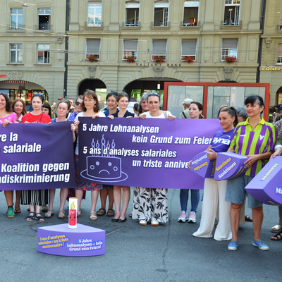 Knapp 30 Frauen aus verschiedenen progressiven Organisationen protestieren in Bern auf dem Bahnhofplatz. Sie stehen vor einem riesigen violetten Kuchenstück aus Styropor - eines von einem ganzen Kuchen, dessen einzelne Stücke die jeweiligen Mängel des Gleichstellungsgesetzes symbolisieren. 