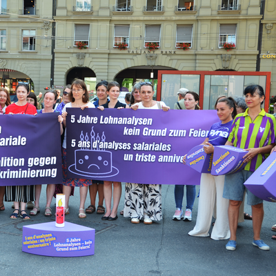 Près de 30 femmes issues de différentes organisations progressistes manifestent à Berne sur la place de la Gare. Elles se tiennent devant un énorme morceau de gâteau violet en polystyrène. Les différentes parts du gâteau symbolisent les lacunes de la loi sur l’égalité. 