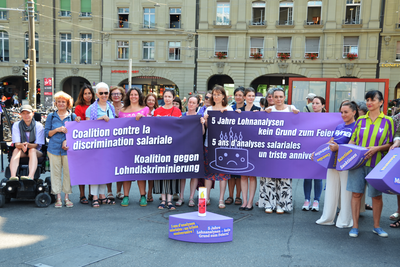 Près de 30 femmes issues de différentes organisations progressistes manifestent à Berne sur la place de la Gare. Elles se tiennent devant un énorme morceau de gâteau violet en polystyrène. Les différentes parts du gâteau symbolisent les lacunes de la loi sur l’égalité. 
