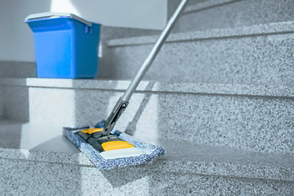 Un balai à serpillère sur une marche d’escalier en pierre claire, avec un seau en plastique bleu à l’arrière-plan. 