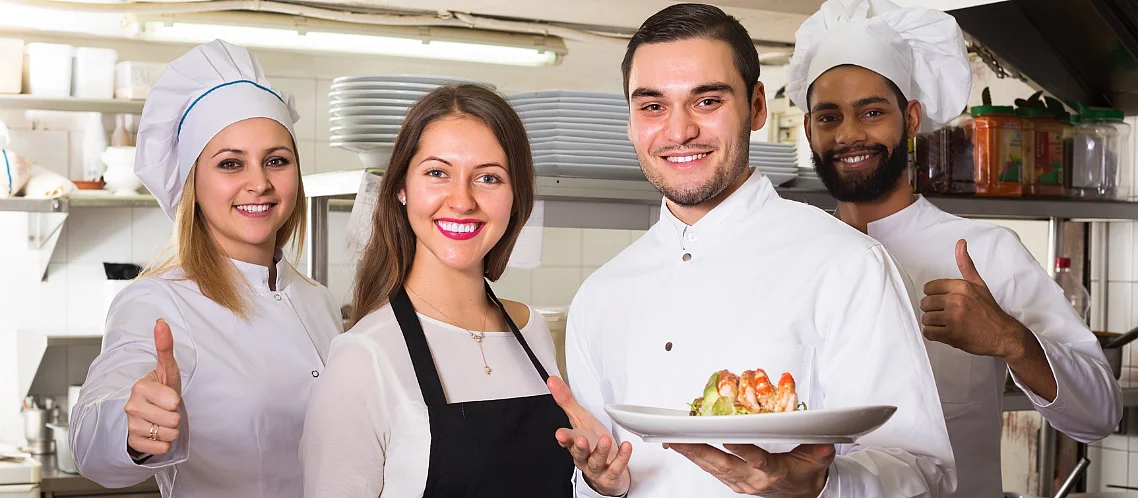 Une cuisinière, deux cuisiniers et une serveuse posent dans une cuisine de restaurant et sourient à la caméra. Deux tiennent des assiettes avec des plats, les autres montrent un pouce en l'air.