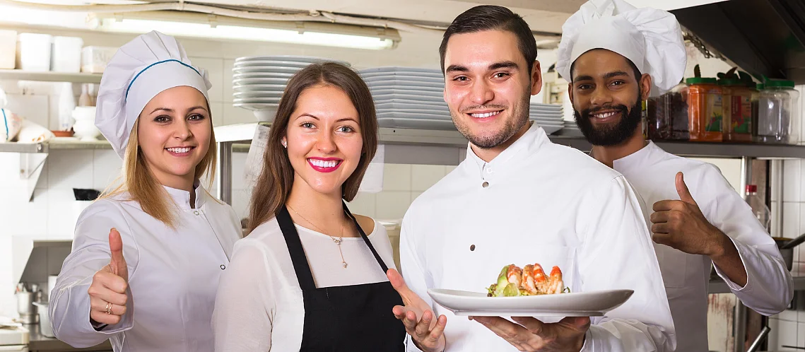 Eine Köchin, zwei Köche und eine Serviceangestellte posieren in einer Restaurantküche und lächeln in die Kamera. Zwei halten Teller mit Gerichten, die anderen zeigen Daumen hoch. 