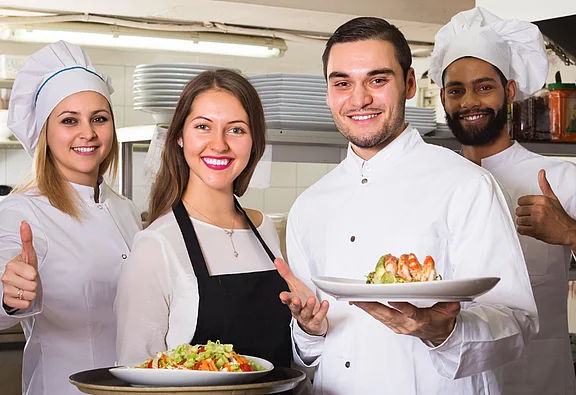 Eine Köchin, zwei Köche und eine Serviceangestellte posieren in einer Restaurantküche und lächeln in die Kamera. Zwei halten Teller mit Gerichten, die anderen zeigen Daumen hoch. 