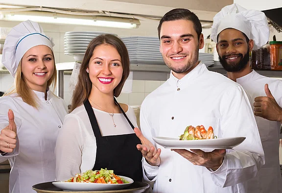  Una cuoca, due cuochi e una cameriera posano in una cucina di ristorante e sorridono alla fotocamera. Due tengono piatti con cibi, gli altri mostrano un pollice in su.