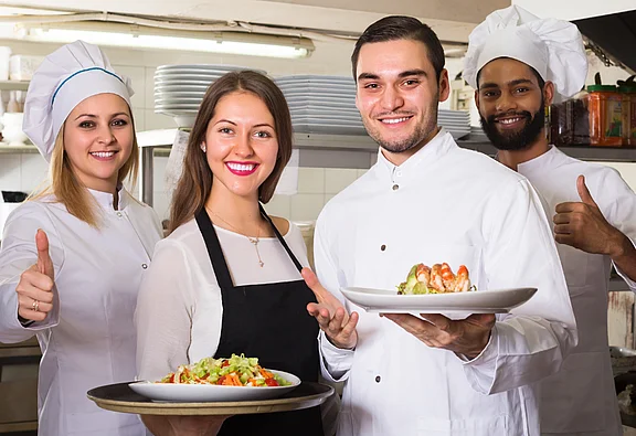 Une cuisinière, deux cuisiniers et une serveuse posent dans une cuisine de restaurant et sourient à la caméra. Deux tiennent des assiettes avec des plats, les autres montrent un pouce en l'air.