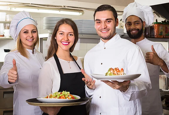  Una cuoca, due cuochi e una cameriera posano in una cucina di ristorante e sorridono alla fotocamera. Due tengono piatti con cibi, gli altri mostrano un pollice in su.