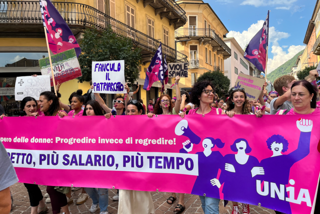 L’immagine mostra una manifestazione con persone che indossano t-shirt rosa e simboli femministi. Una persona in primo piano indossa una t-shirt con lo slogan «Rispetto, più salario, più tempo!». I partecipanti reggono fumogeni colorati che producono fumo viola. Sullo sfondo è visibile un ambiente urbano con edifici e una torre dell’orologio. L’atmosfera è determinata e combattiva.