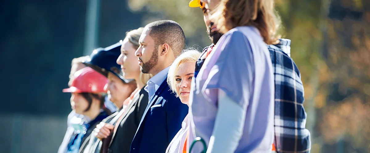 La photo montre une vue latérale d'une série de six personnes debout représentant des collaborateurs de différentes branches d'Unia. Chaque personne porte des vêtements professionnels représentant son secteur, comme des casques et des combinaisons.