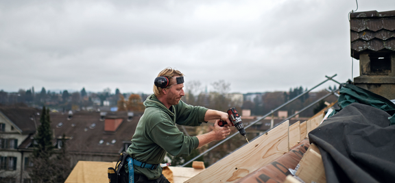 Un ouvrier avec protection auditive et une ceinture porte-outils utilise une visseuse à accus sur une charpente en milieu urbain.