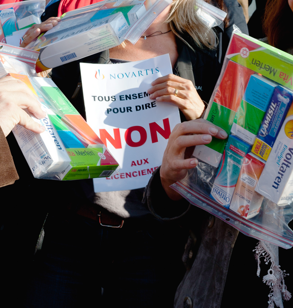 Action contre des licenciements chez Novartis: des personnes montrent des boîtes de médicaments (Voltaren, Otrivin) et une pancarte sur laquelle est inscrit « Tous ensemble pour dire NON aux licenciements ».