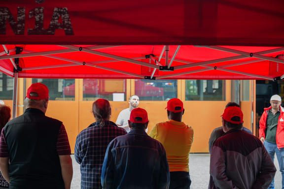 Im Vordergrund stehen Streikende mit dem Rücken zur Kamera und hören einem Gewerkschaftssekretär zu. Sie stehen in einem Zelt der Gewerkschaft Unia vor einem Gebäude der Firma Samvaz SA. 