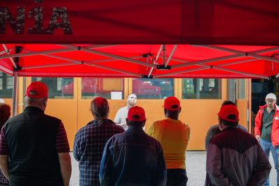 Au premier plan, des grévistes de dos écoutent un secrétaire syndical. Ils se trouvent sous une tente Unia, devant un bâtiment de l’entreprise Samvaz SA. 