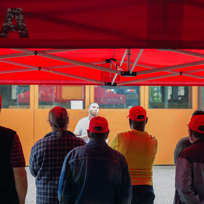 Au premier plan, des grévistes de dos écoutent un secrétaire syndical. Ils se trouvent sous une tente Unia, devant un bâtiment de l’entreprise Samvaz SA. 