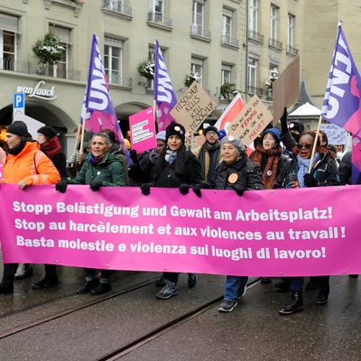 Eine Gruppe von Menschen demonstriert in der Berner Altstadt. Sie gehen auf einer Strasse mit Tramgleisen und tragen mehrere violette Unia Fahnen. Vorne halten sie ein grosses pinkes Unia-Banner mit Forderungen gegen Belästigung und Gewalt am Arbeitsplatz in Deutsch, Französisch und Italienisch. Weitere Teilnehmende tragen Plakate mit politischen Botschaften. Es liegt Schnee am Strassenrand, und im Hintergrund stehen mehrstöckige Gebäude mit Balkonen. Die Stimmung wirkt engagiert und kämpferisch.