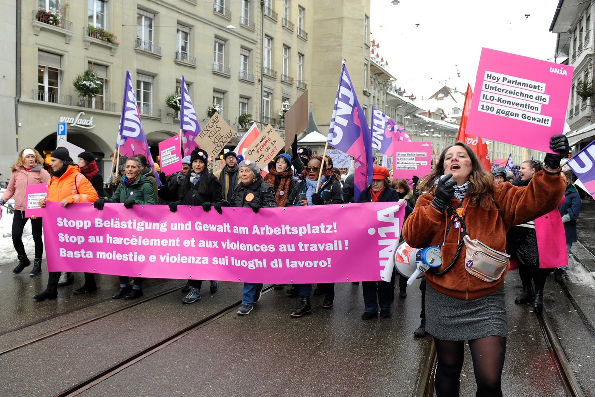 Eine Gruppe von Menschen demonstriert in der Berner Altstadt. Sie gehen auf einer Strasse mit Tramgleisen und tragen mehrere violette Unia Fahnen. Vorne halten sie ein grosses pinkes Unia-Banner mit Forderungen gegen Belästigung und Gewalt am Arbeitsplatz in Deutsch, Französisch und Italienisch. Weitere Teilnehmende tragen Plakate mit politischen Botschaften. Es liegt Schnee am Strassenrand, und im Hintergrund stehen mehrstöckige Gebäude mit Balkonen. Die Stimmung wirkt engagiert und kämpferisch.