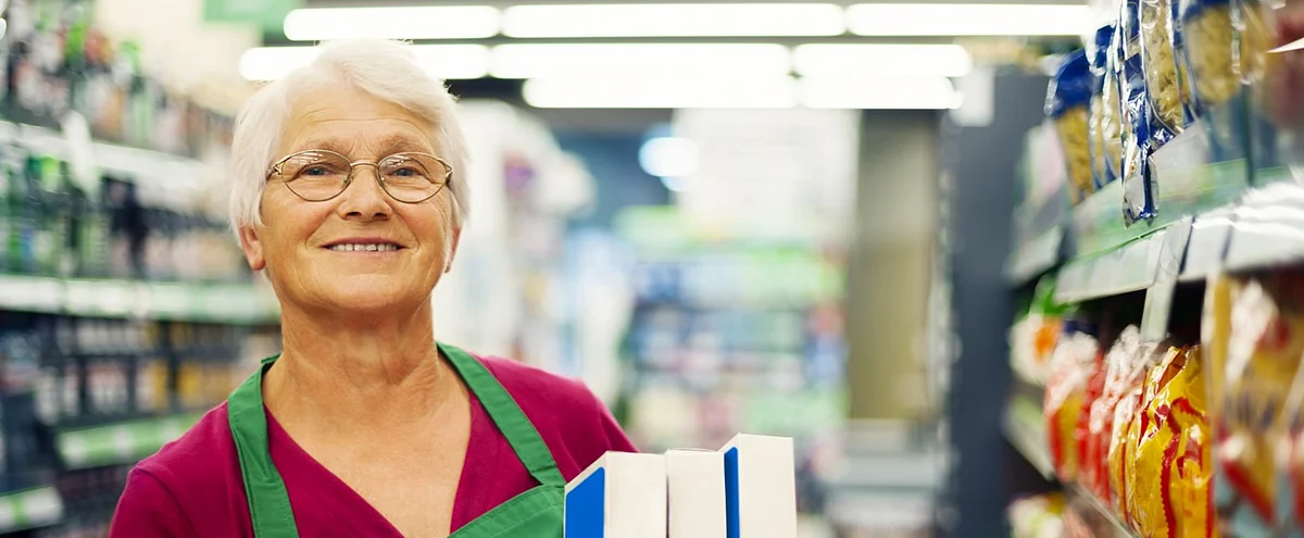 Une vendeuse âgée aux cheveux blancs et portant des lunettes sourit à la caméra. Elle porte un tablier vert et tient des produits dans une allée de supermarché. En arrière-plan, les étagères avec des aliments sont visibles de manière floue.