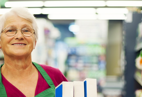 Une vendeuse âgée aux cheveux blancs et portant des lunettes sourit à la caméra. Elle porte un tablier vert et tient des produits dans une allée de supermarché. En arrière-plan, les étagères avec des aliments sont visibles de manière floue.