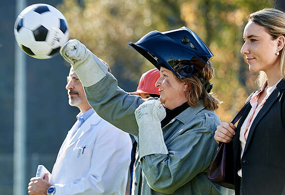 Une femme avec un casque et des gants de soudeur lance une balle. À coté d'elle: un homme en tablier de laboratoire et une femme en costume de ville.