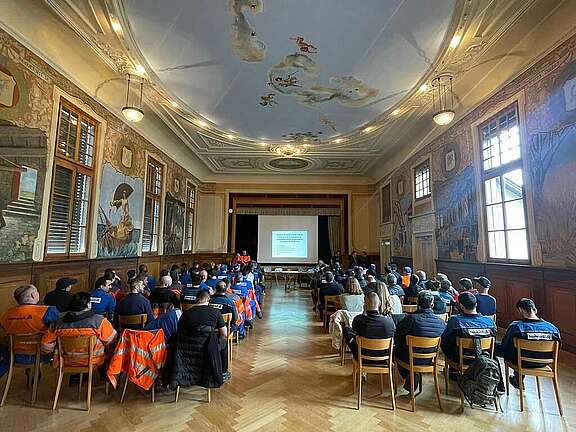 Arbeiter und Arbeiterinnen von Vetropack sitzen auf Stühlen in einem grossen Versammlungssaal