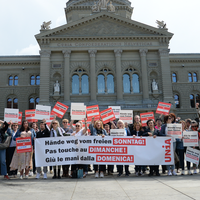 Un groupe d’environ 50 personnes portant des panneaux et une banderole devant le Palais fédéral à Berne. Sur la banderole, on peut y lire en trois langues «Pas touche au congé du dimanche!».