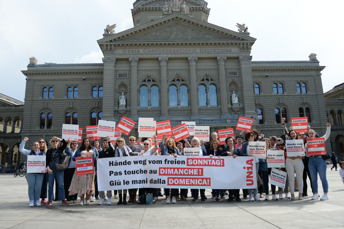 Eine Gruppe von ca. 50 Menschen mit Schildern und einem Transparent vor dem Bundeshaus in Bern. Auf dem Transparent ist in drei Sprachen «Hände weg vom freien Sonntag!» zu lesen.