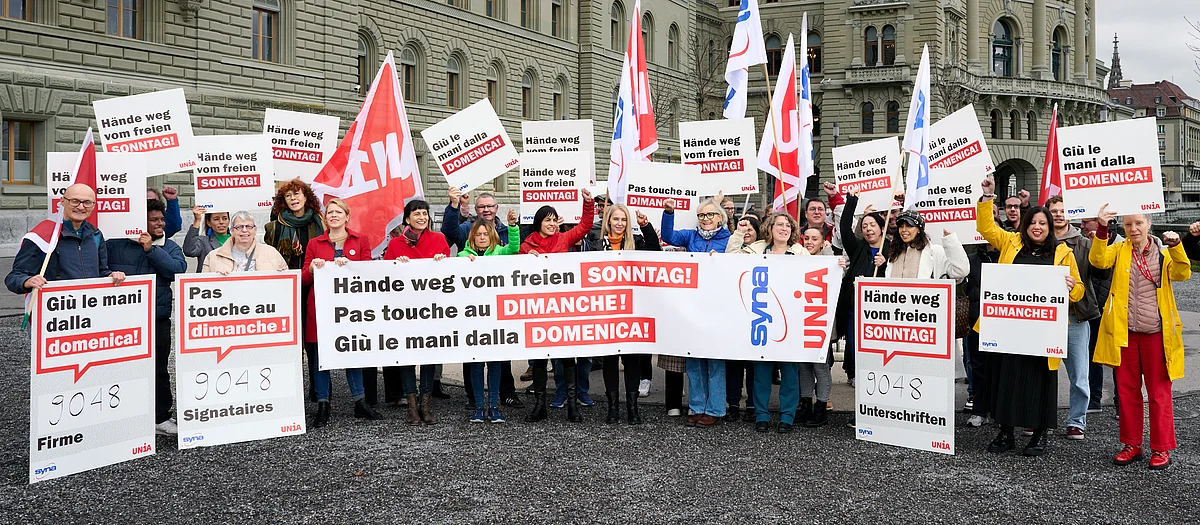 Menschen halten ein Transparent und Plakate in die Luft, auf denen «Hände weg vom freien Sonntag» in Deutsch, Französisch und Italienisch steht.