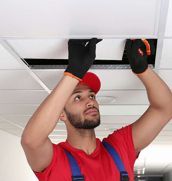 Ein Arbeiter mit roten Arbeitskleidern und Handschuhen installiert eine Deckenplatte in einer abgehängten Rasterdecke und hält dabei eine Zange in der Hand.