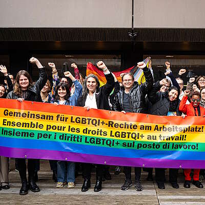 Un gruppo di persone tiene uno striscione arcobaleno con la scritta «Insieme per i diritti LGBTQI+ sul posto di lavoro!» e alza il pugno con determinazione.