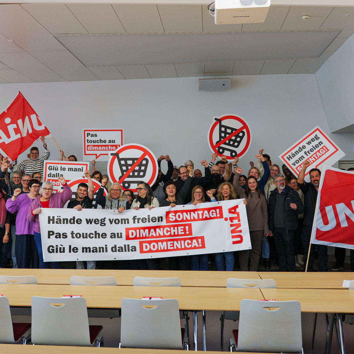 Un gruppo di persone si riunisce dietro uno striscione con la scritta «Giù le mani dalla domenica libera». Tengono in mano cartelli e sventolano bandiere Unia.