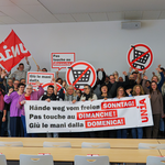 Eine Gruppe von Menschen versammelt sich hinter einem Transparent, auf dem steht «Hände weg vom freien Sonntag». Sie halten Schilder und schwenken Unia-Fahnen.