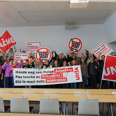 Un gruppo di persone si riunisce dietro uno striscione con la scritta «Giù le mani dalla domenica libera». Tengono in mano cartelli e sventolano bandiere Unia.