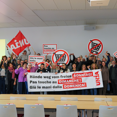 Un groupe de personnes rassemblées derrière une banderole «Pas touche au dimanche!» brandissent des pancartes et agitent des drapeaux Unia.