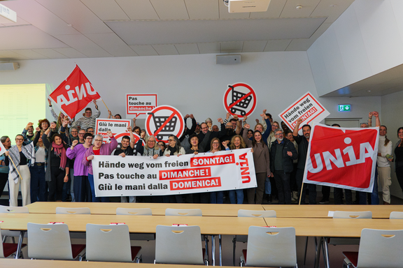Eine Gruppe von Menschen versammelt sich hinter einem Transparent, auf dem steht «Hände weg vom freien Sonntag». Sie halten Schilder und schwenken Unia-Fahnen.