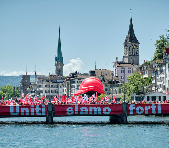 Manifestation de la construction à Zurich avec un grand casque rouge comme symbole sur un pont. En arrière-plan, on aperçoit les tours emblématiques du Grossmünster et du Fraumünster. Sur le pont, une banderole porte l'inscription 'Uniti siamo forti' (Ensemble, nous sommes forts), et de nombreux participants brandissent des drapeaux rouges.