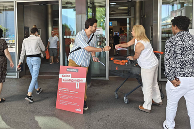 Protesta di prima mattina davanti all'ingresso della Migros contro i tagli ai posti di lavoro. Distribuiscono volantini e spille.