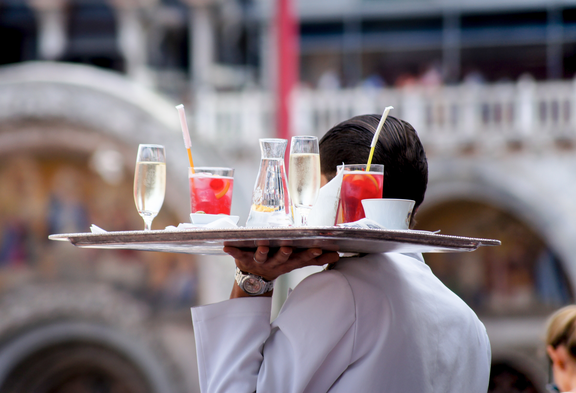 Da dietro arriva un cameriere con un vassoio pieno di bevande.
