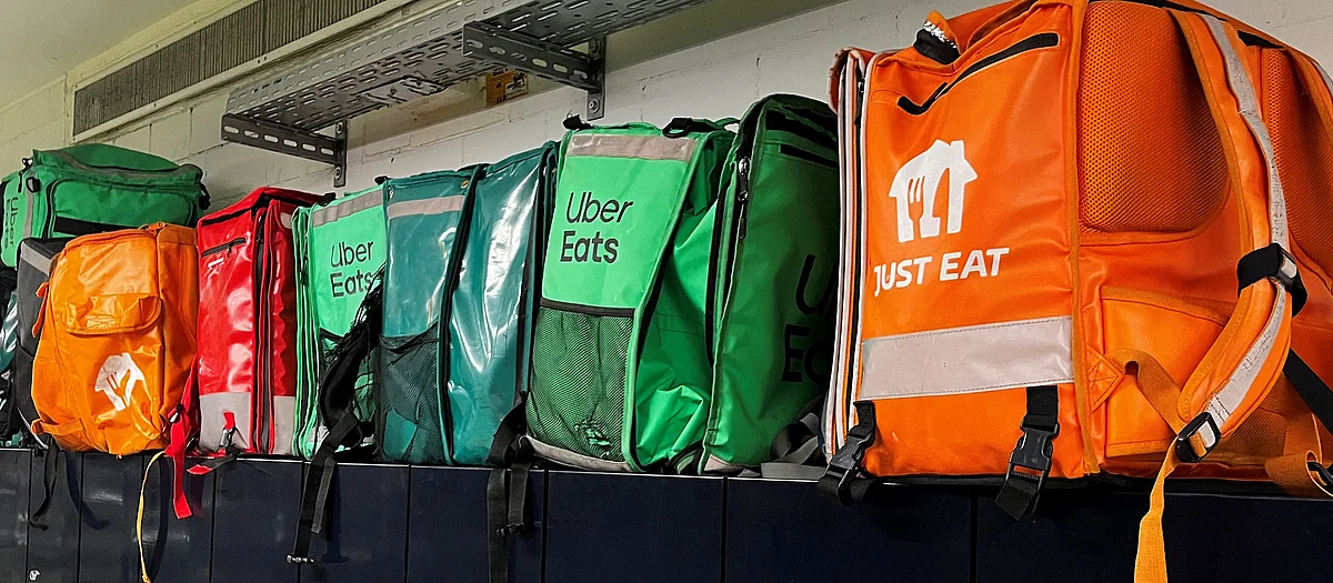 Sacs à dos de livraison verts d’Uber Eats et orange de Just Eat alignés côte à côte sur une étagère contre un mur blanc.