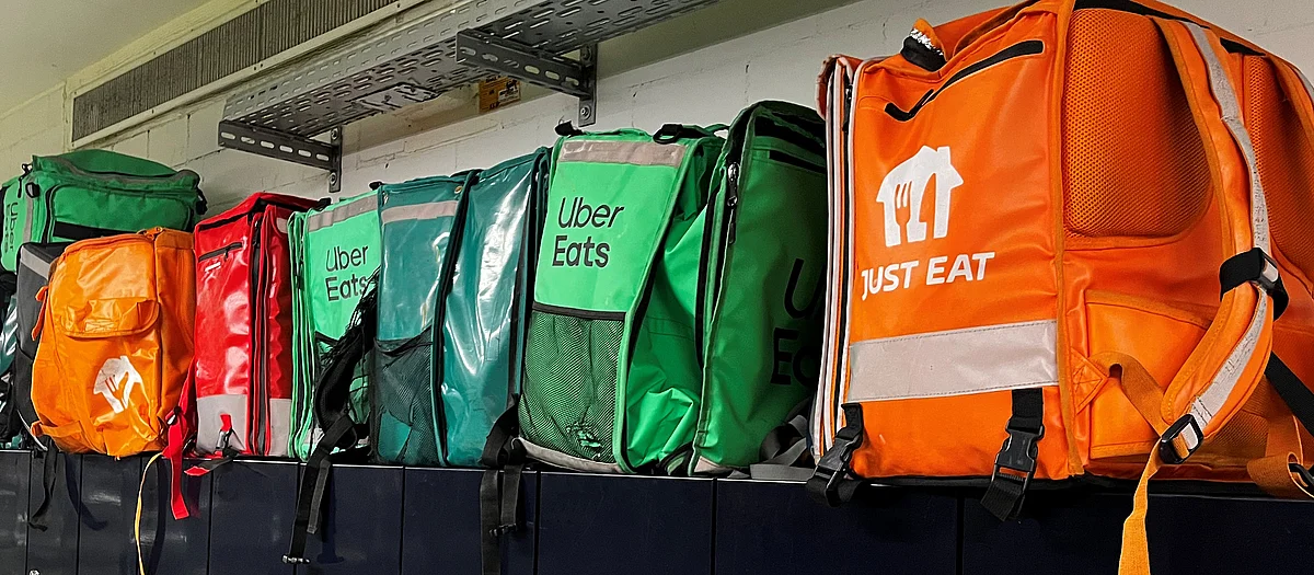 Grüne Uber-Eats- und orange Just-Eat-Lieferrucksäcke stehen dicht aufgereiht auf einem Regal vor einer weissen Wand.