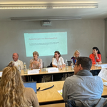 Photo de la conférence de presse de l’Alliance pour le dimanche. Les intervenant-e-s sont assis à une table de conférence. On peut voir devant eux des plaquettes avec leurs noms et, derrière eux, un écran où sont projetés les contenus de l’étude.