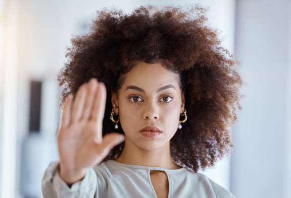 Una giovane donna con i capelli ricci e un'espressione seria sul volto tiene la mano davanti a sé con le dita divaricate, segnalando che deve fermarsi.