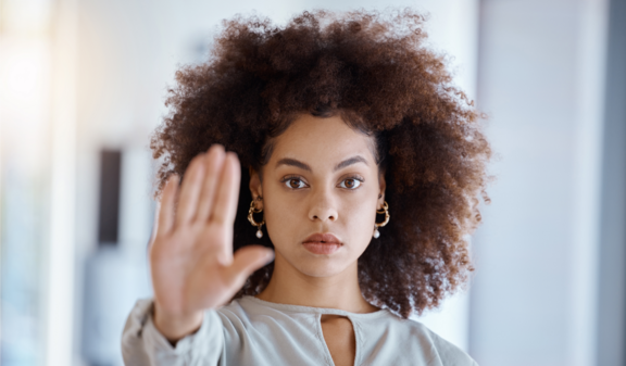 Une jeune femme aux cheveux bouclés et à l'expression grave tient sa main devant elle, les doigts écartés, en signe d'arrêt.