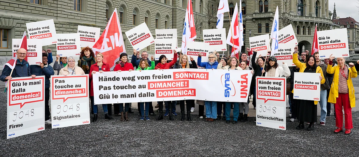 Devant le Palais fédéral, des personnes manifestent contre le travail dominical avec des pancartes multilingues, des banderoles et des drapeaux Unia, et réclament la protection du dimanche.
