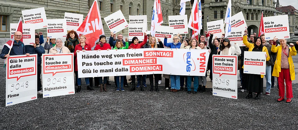 Davanti al Palazzo federale, un gruppo di persone protestano con cartelli multilingue, striscioni e bandiere Unia contro il lavoro domenicale e chiedono la tutela della domenica libera.