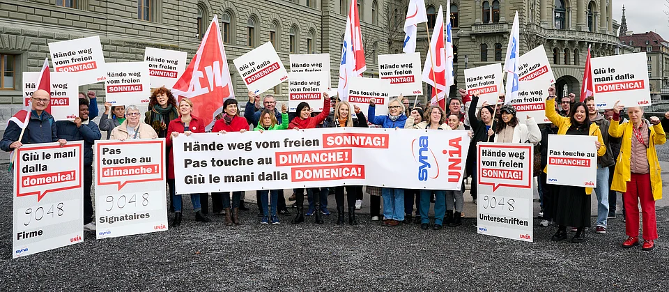 Devant le Palais fédéral, des personnes manifestent contre le travail dominical avec des pancartes multilingues, des banderoles et des drapeaux Unia, et réclament la protection du dimanche.