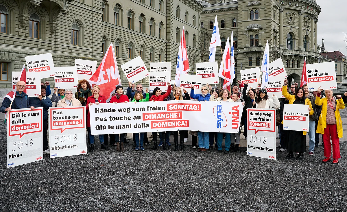 Davanti al Palazzo federale, un gruppo di persone protestano con cartelli multilingue, striscioni e bandiere Unia contro il lavoro domenicale e chiedono la tutela della domenica libera.
