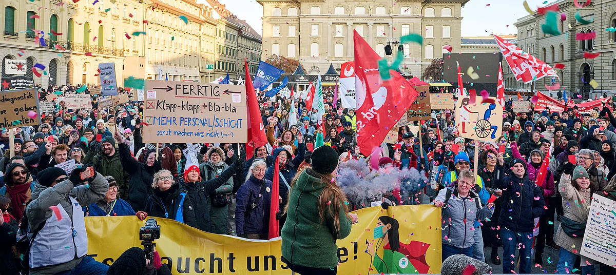  Pflegepersonal demonstriert mit Transparenten, Fahnen und Schildern auf dem Bundesplatz in Bern, im Vordergrund sind Botschaften zu Arbeitsbedingungen und Gleichstellung sichtbar.