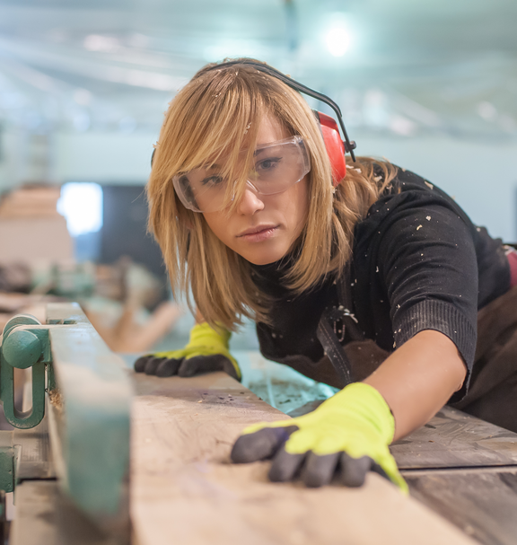Una persona lavora una tavola di legno utilizzando una macchina fissa in una falegnameria, indossando indumenti di protezione.